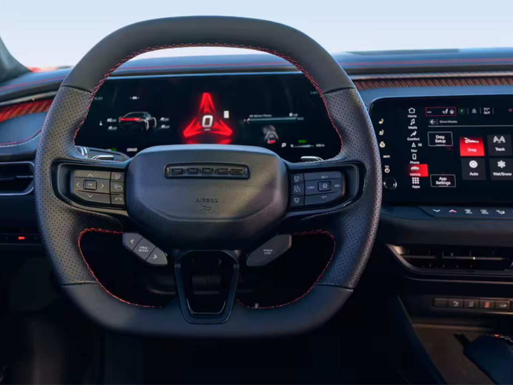 The dashboard and steering wheel of the Dodge Charger Daytona. The steering wheel has a flat bottom and mounted controls. The screens show red accents and buttons to tap for settings and entertainment.